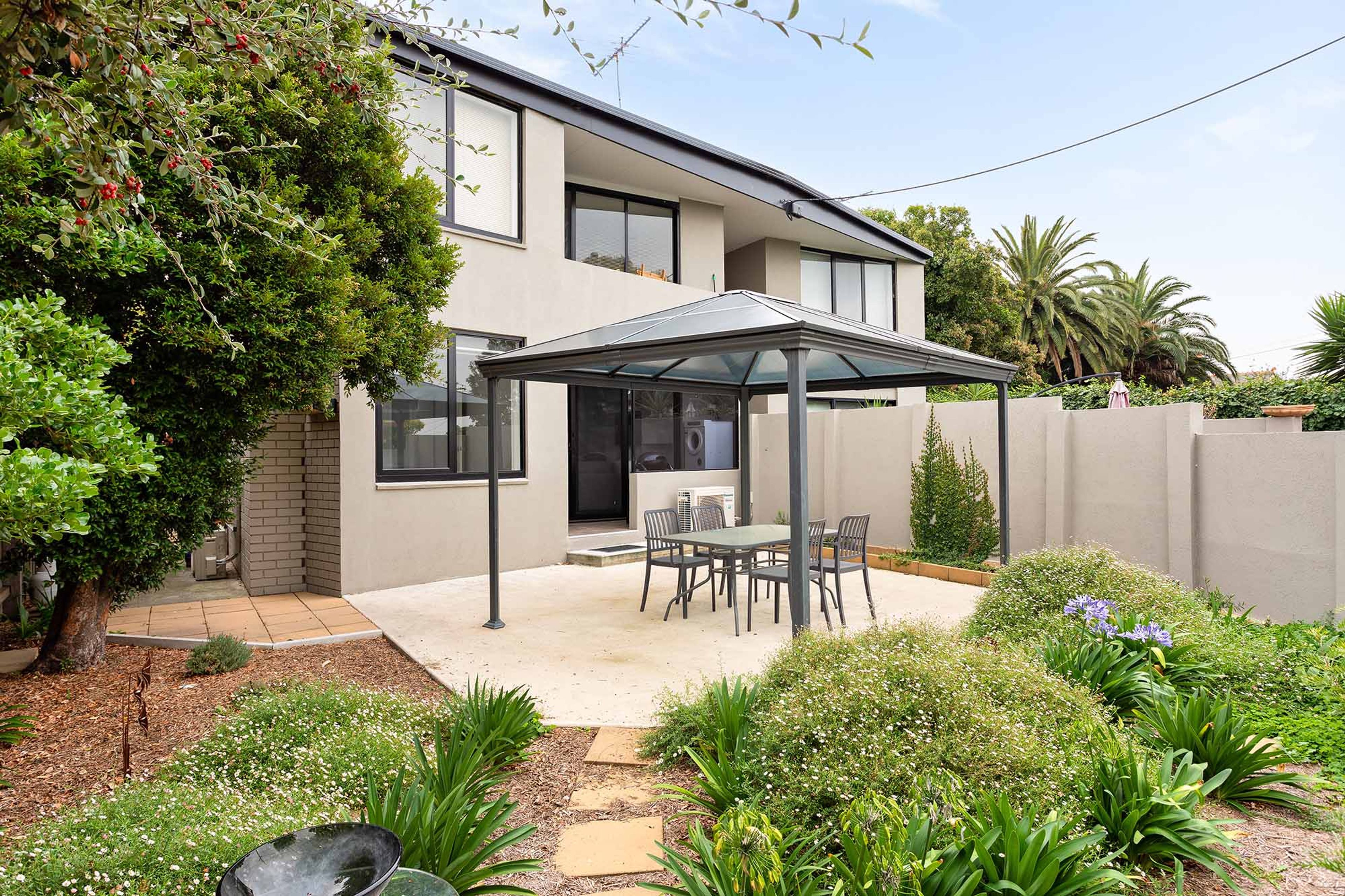 A modern two-story house with a covered patio, a brick wall, and a garden with various plants and flowers.