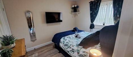 A bedroom with a bed, a TV, a window with curtains, a bedside table, and a lamp.