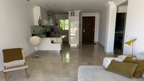 A modern living room with a grey sofa, a white armchair, and a coffee table. There is a kitchen area in the background with a glass island and pendant lights. - Apartment Aloha Hill Club Marbella (Marbella)