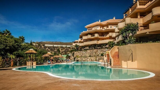 A swimming pool with a curved edge, surrounded by a stone patio. There are lounge chairs and umbrellas in the background. - Apartment Aloha Hill Club Marbella (Marbella)