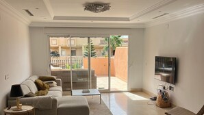 A spacious living room with a large sofa, a TV mounted on the wall, and a sliding glass door leading to an outdoor area. - Apartment Aloha Hill Club Marbella (Marbella)