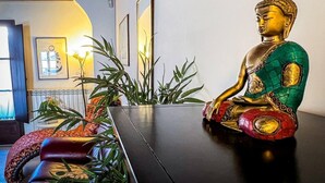 En Buddha-staty står på ett bord i ett rum med ett träpanelat tak, inramade konstverk hänger på väggen och en växt syns i förgrunden.