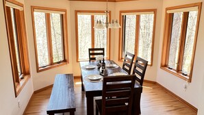 Comedor con mesa de madera, sillas y banco. Cuenta con ventanas amplias con marcos de madera y una araña.