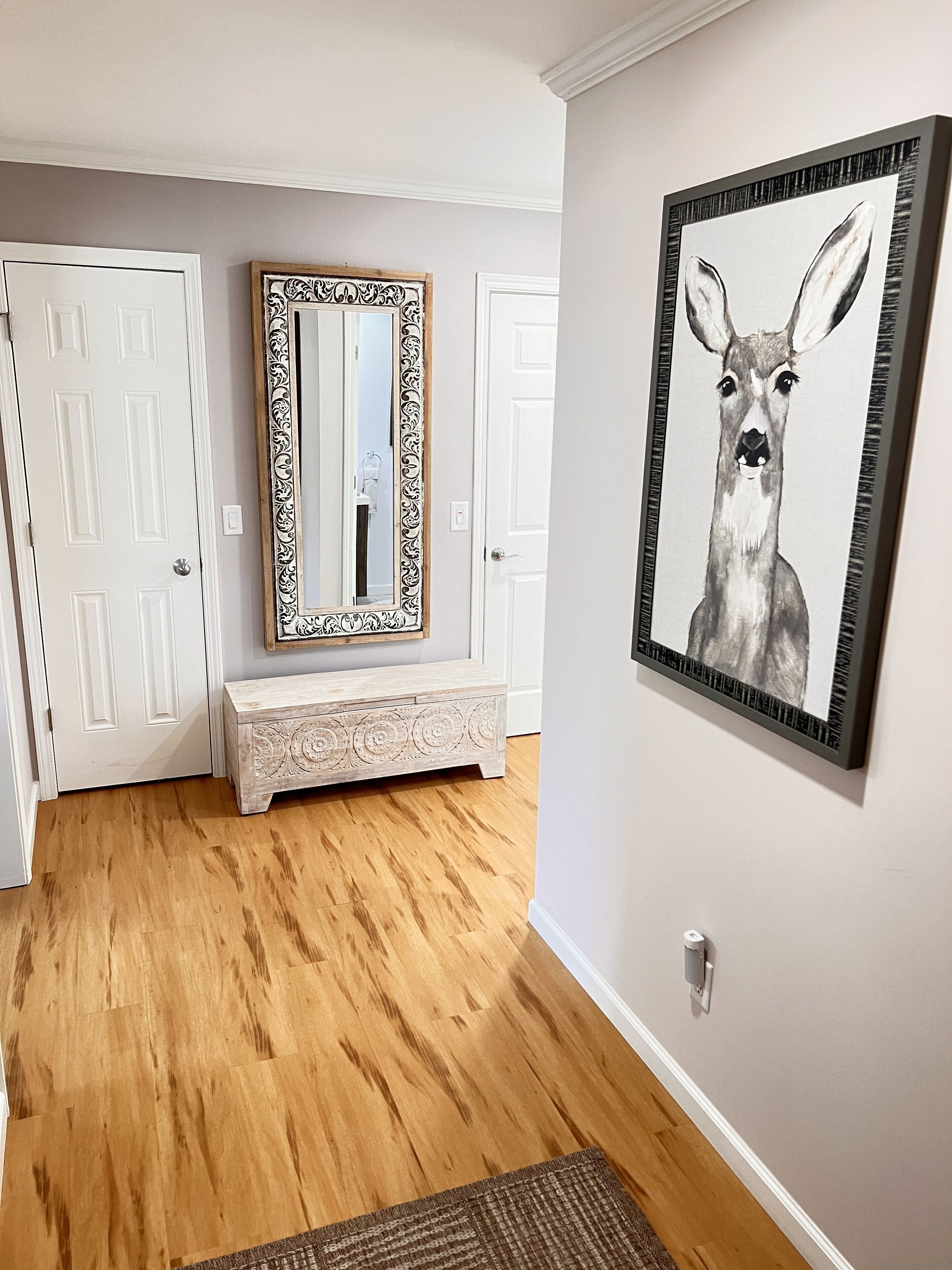 Un couloir doté d’un plancher en bois, de portes blanches, d’un grand miroir encadré d’un cadre orné, d’un banc et d’une œuvre d’art encadrée représentant un cerf.
