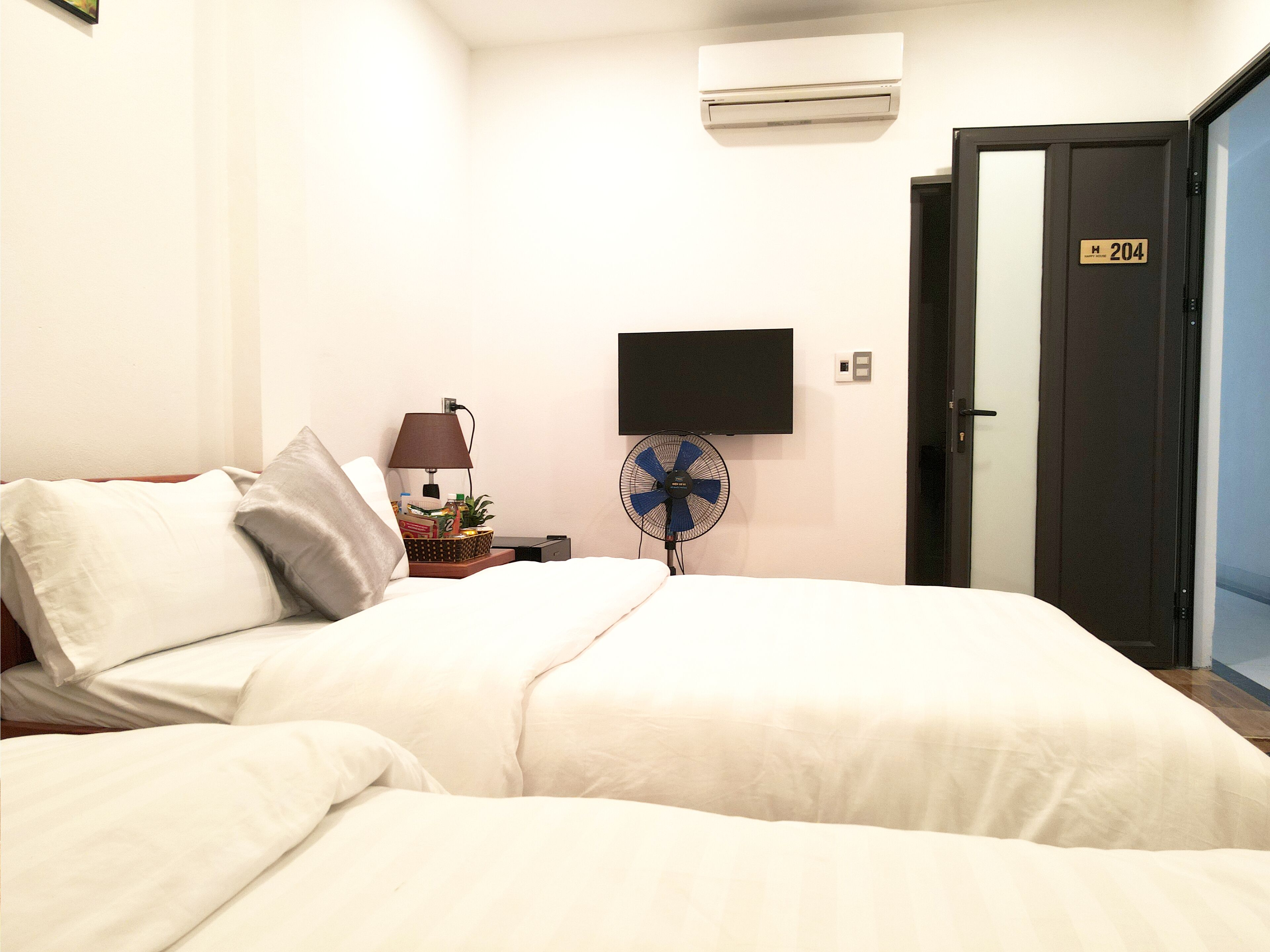 Une chambre d’hôtel comprenant un lit, une table de chevet avec une lampe, un téléviseur fixé au mur, un climatiseur et une porte avec le numéro de la chambre.
