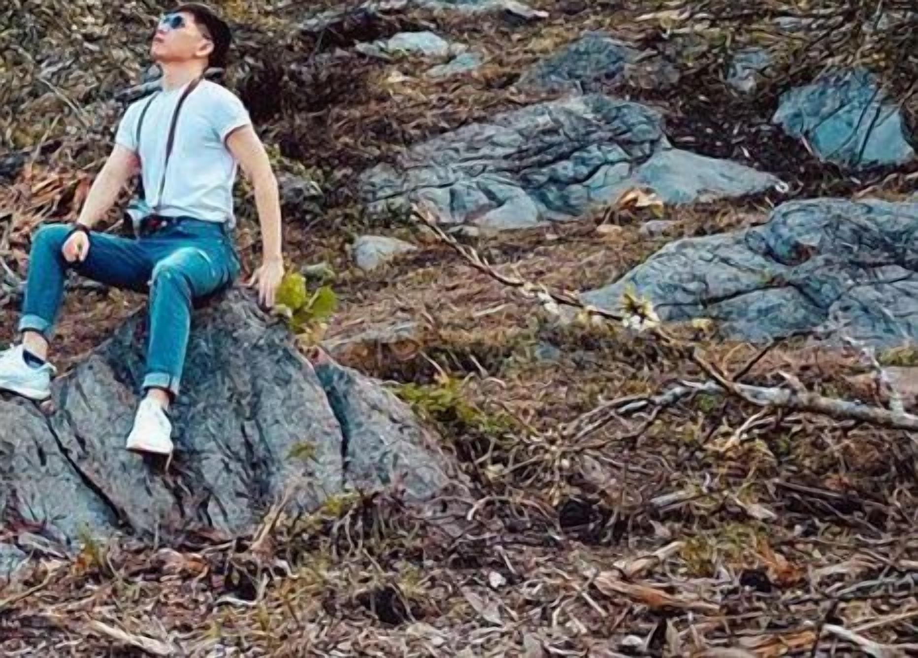 A person sitting on a rock in a natural setting with trees and foliage around.