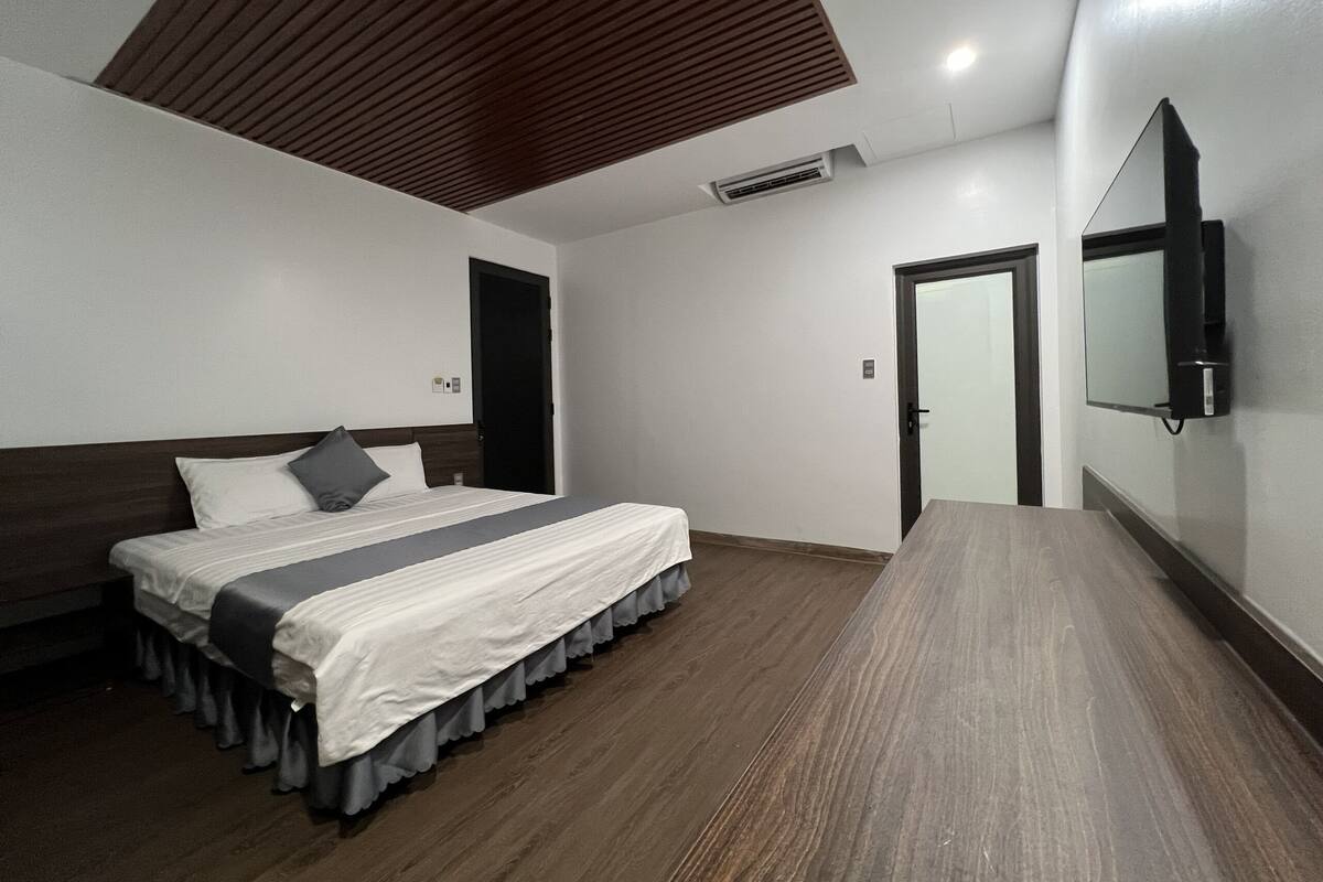Quarto com cama, teto de madeira, televisão na parede e uma porta.