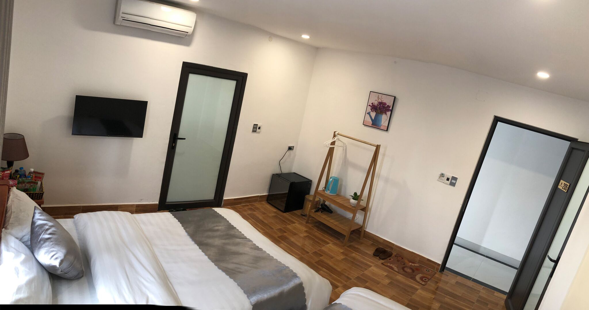 A bedroom with a bed, a TV mounted on the wall, a door, a small table with a plant, and a wall-mounted air conditioner.