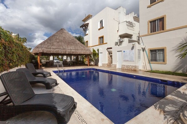 Una piscina con sillas de descanso, una estructura de techo de paja y un edificio con varios balcones.