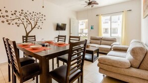 Un comedor con una mesa de madera, sillas y un televisor montado en la pared. Se observa un sofá y una ventana con cortinas al fondo.