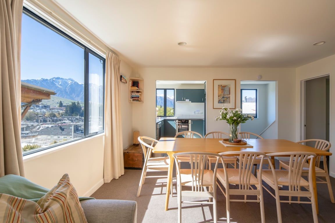 Un área de comedor con una mesa y sillas, vistas de la ciudad y las montañas a través de un ventanal grande, y una kitchenette al fondo.