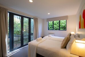Habitación de hotel con un amplio ventanal corredera que da a un jardín, una cama con almohadas, una mesita de noche y una ventana con vistas a vegetación.