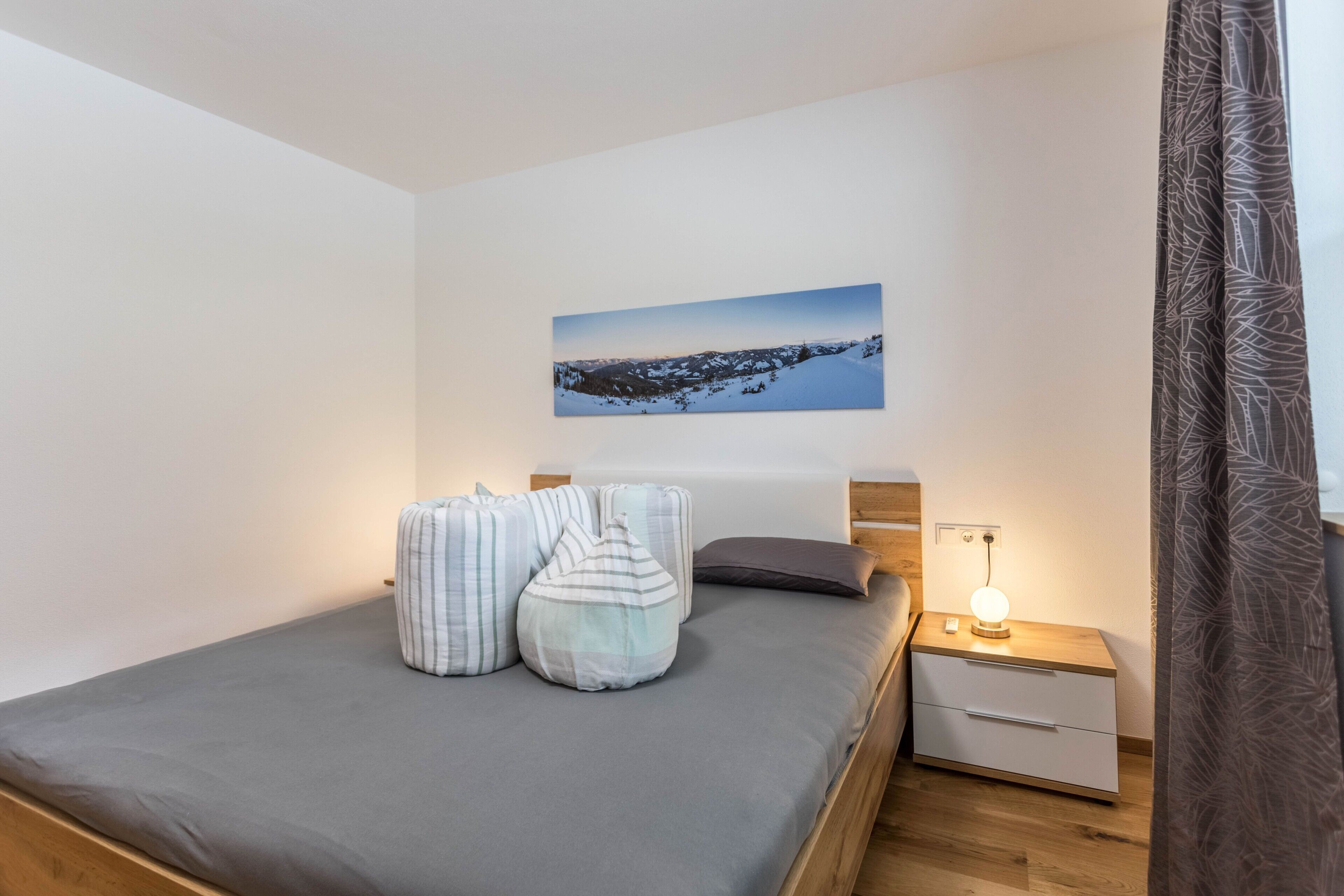 A bedroom with a bed, pillows, a nightstand with a lamp, a painting on the wall, and a wooden headboard.