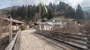 Un pont en bois mène à un village où se trouvent des maisons traditionnelles, un drapeau et, en arrière-plan, une colline boisée.