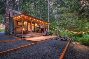 A cabin with a wooden deck, string lights, and a view of the forest. - Li'l Finch Mt. Rainier tiny cabin with covered Hot tub and steam sauna (Ashford)