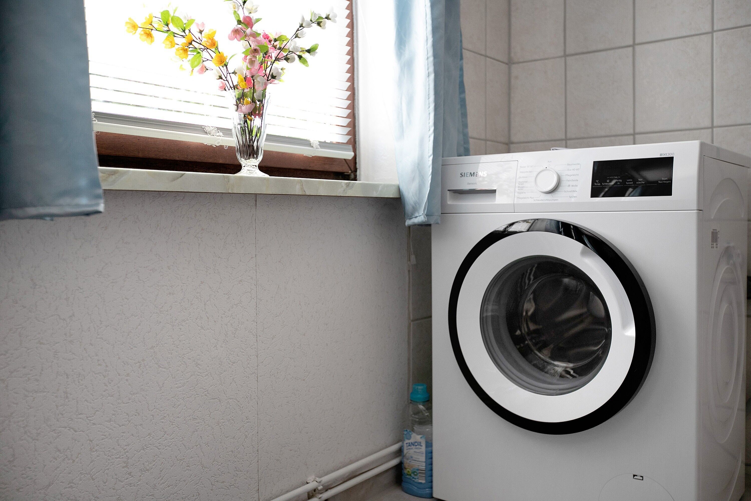 A white washing machine in a bathroom with a window, a vase with flowers, and a bottle of detergent.