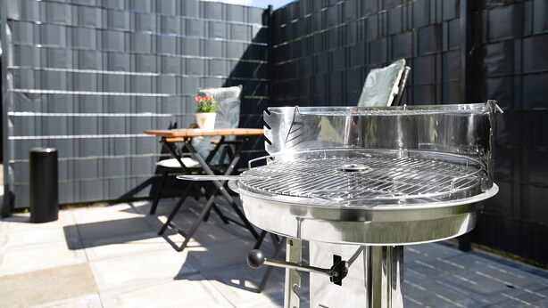 Ein Grill aus Edelstahl mit Deckel, ein Tisch, Stühle und ein Blumentopf im Garten.