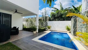 A modern outdoor pool area with a ceiling fan, a seating area, and a stone wall with tropical plants. - Lujo en el Centro de Bonao Piscina Privada King Bed (Bonao)