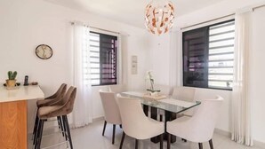 A modern dining area with a glass table, white chairs, and a wooden bar counter. - Lujo en el Centro de Bonao Piscina Privada King Bed (Bonao)