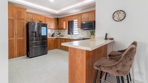 A modern kitchen with wooden cabinets, a stainless steel refrigerator, a microwave, and a bar area with two cushioned stools. - Lujo en el Centro de Bonao Piscina Privada King Bed (Bonao)