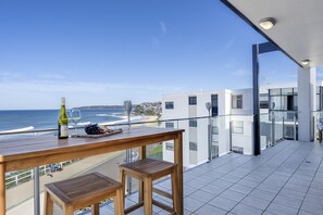 Un balcon avec une table en bois et deux tabourets, donnant sur une plage et l’océan.