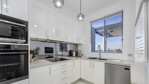 A modern kitchen with white cabinets, stainless steel appliances, and a large window offering a view of the sea.
