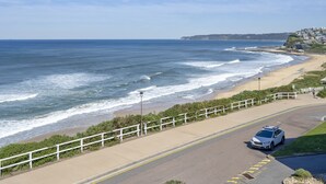 A coastal road with a white car, a beach, and a white fence along the road.