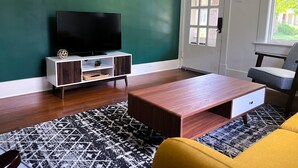 Un soggiorno con una TV a schermo piatto su un mobiletto bianco e in legno scuro, un tappeto decorato e un divano giallo.