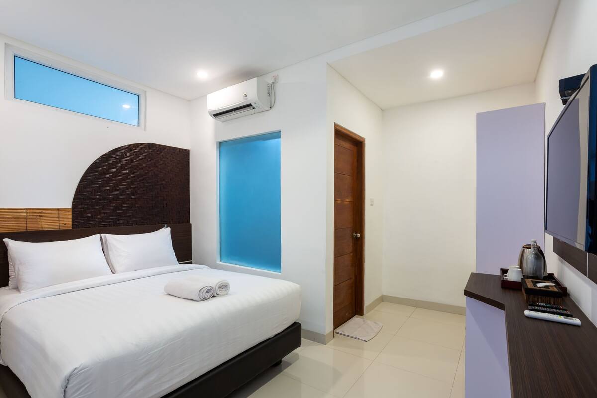 A modern hotel room with a large bed, a flat-screen TV, a wooden headboard, a small desk, and a window with a view.