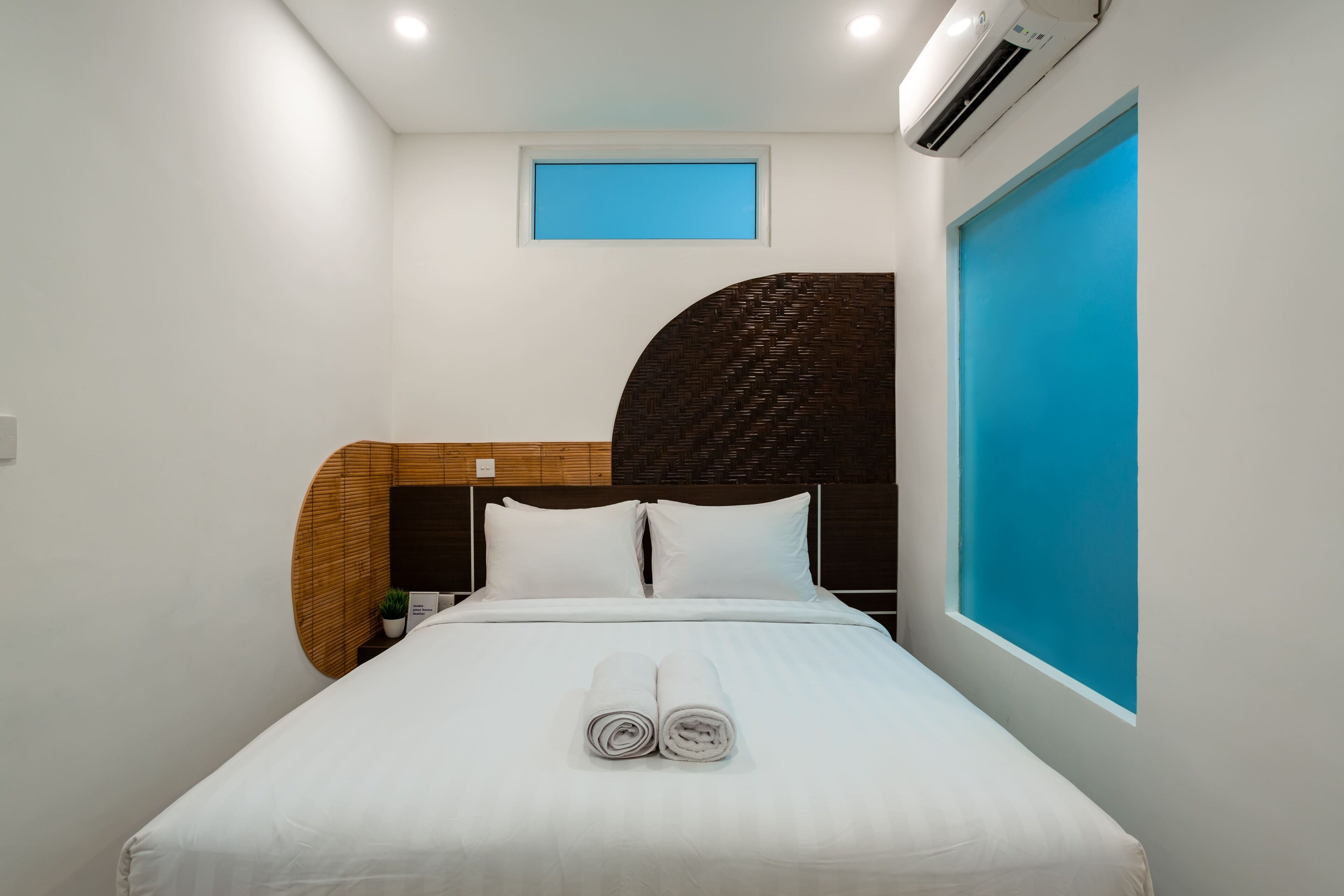 A modern hotel room with a neatly made bed, a small window with a blue view, and a wall-mounted air conditioner.