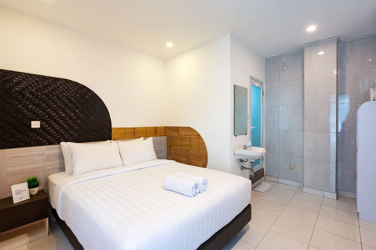 A modern hotel room with a large bed, a bedside table with a plant, a bathroom with a sink and mirror, and a shower area.