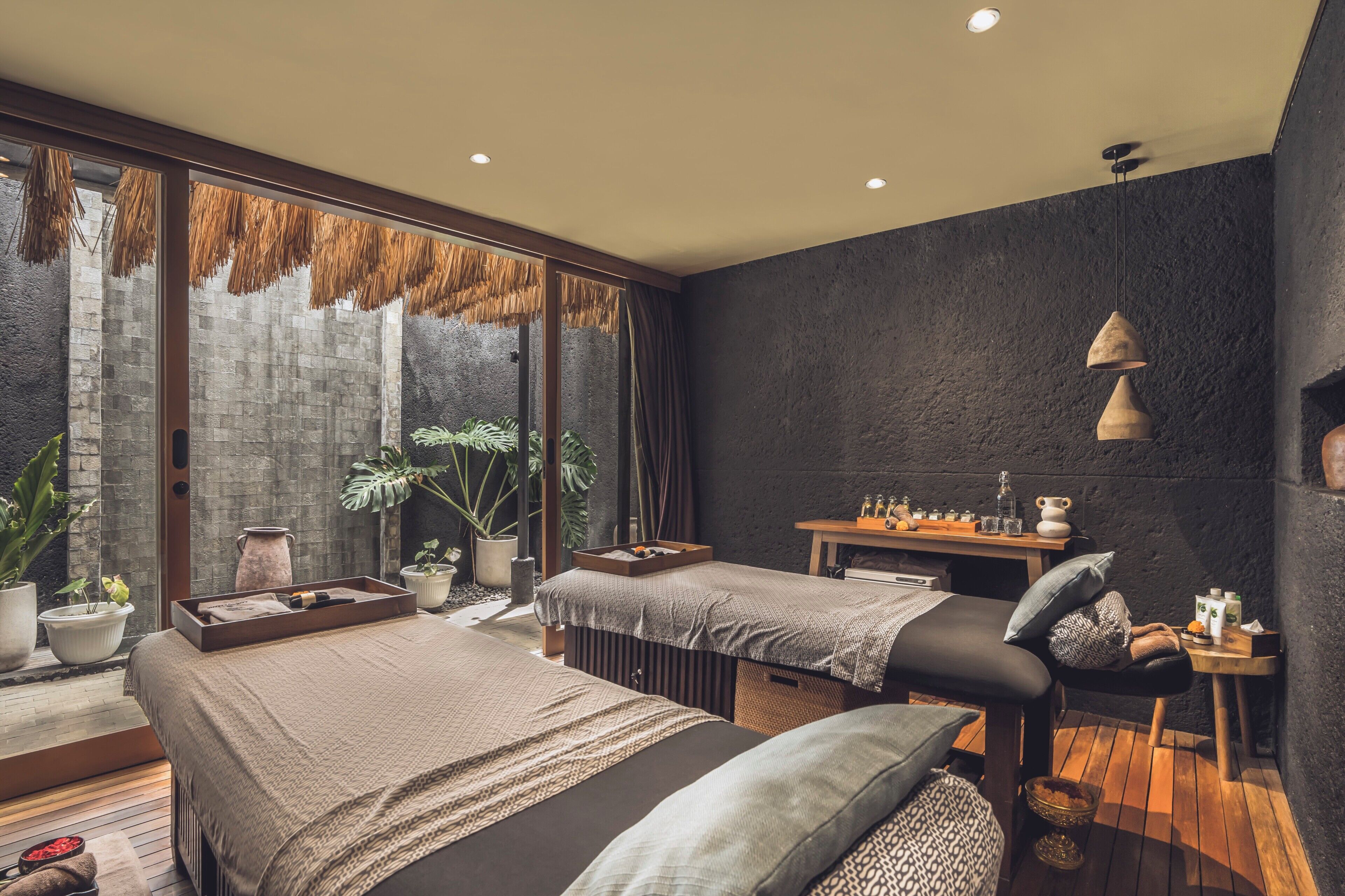 Una habitación moderna con dos camas, una camilla de masaje, un escritorio pequeño y ventanas amplias con vistas a un muro de piedra y plantas.