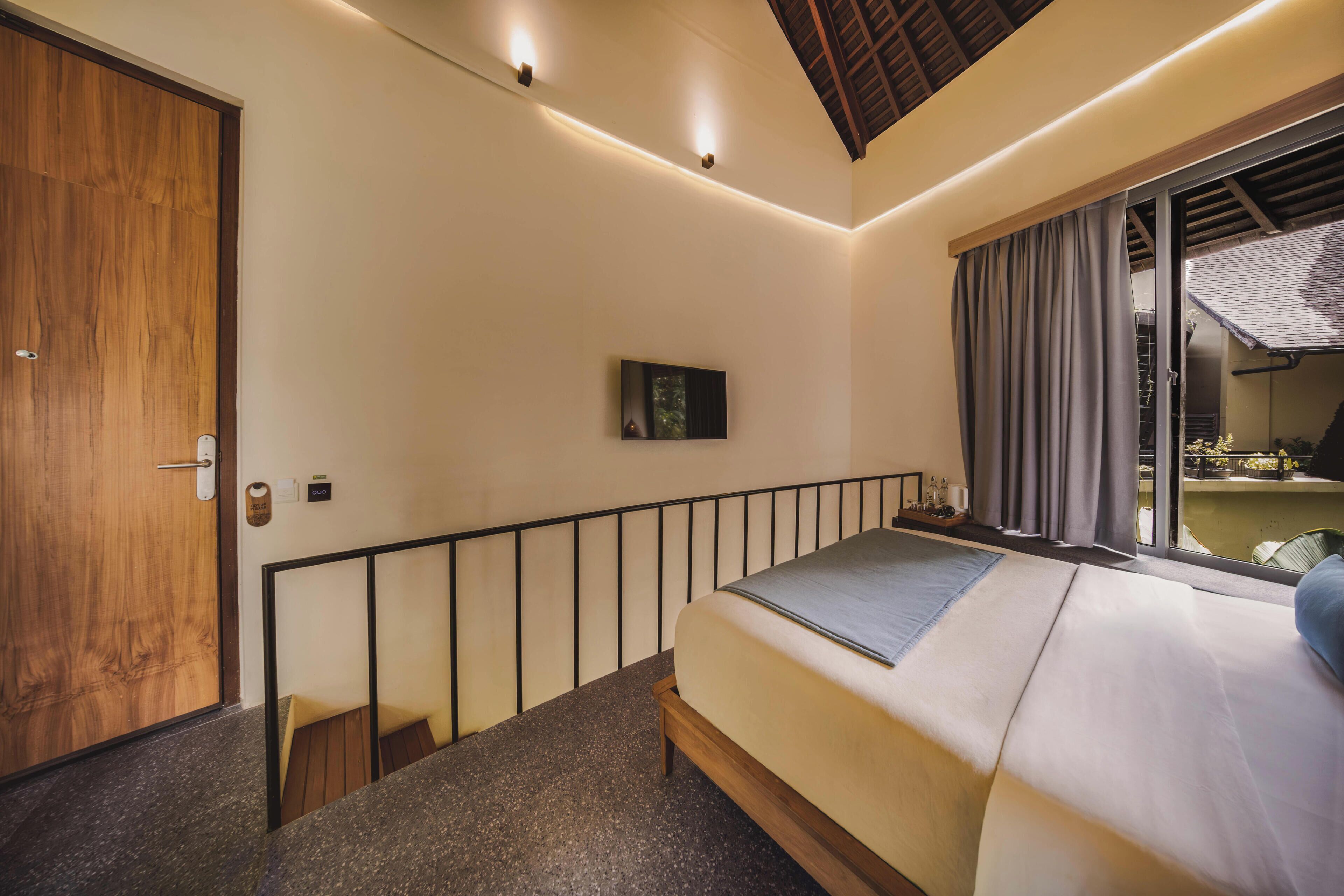 Une chambre à coucher avec un lit, une porte en bois, une petite table, un téléviseur et un balcon avec des rideaux.
