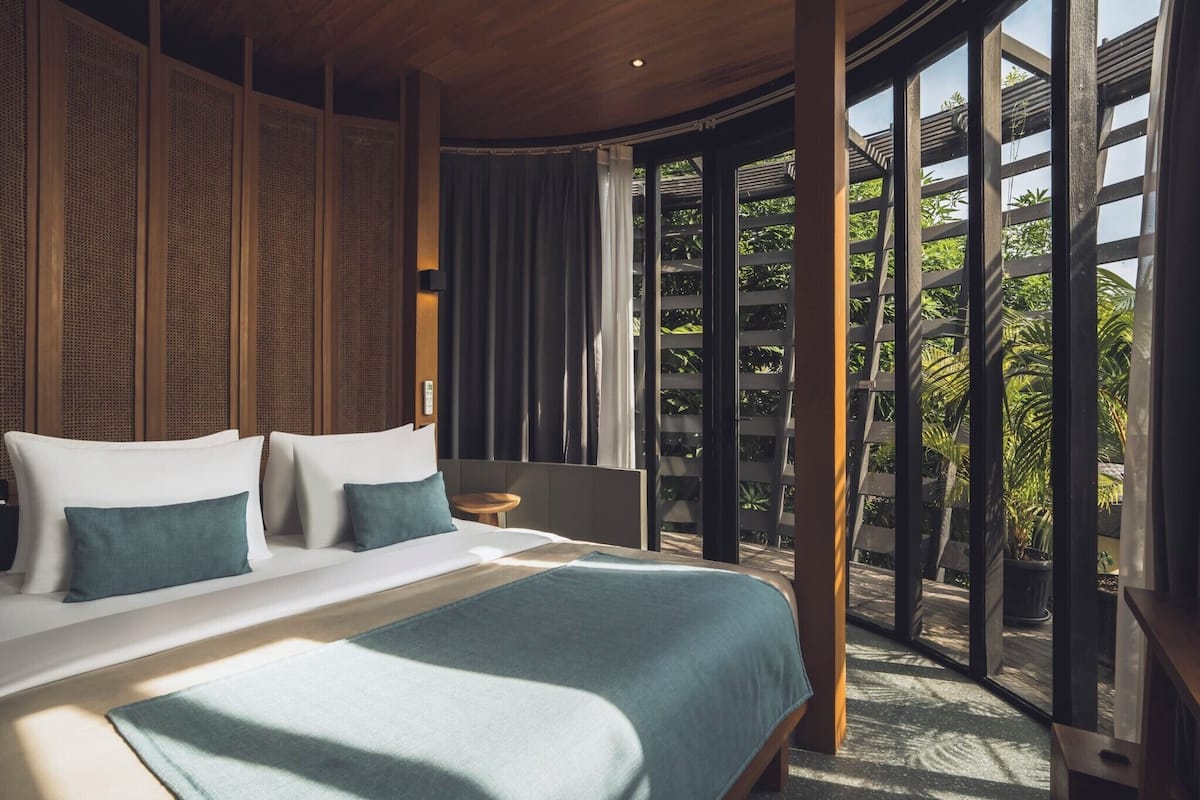 Ein modernes Schlafzimmer mit einem großen Bett, einem kleinen Nachttisch und Blick auf Grün durch große Fenster.
