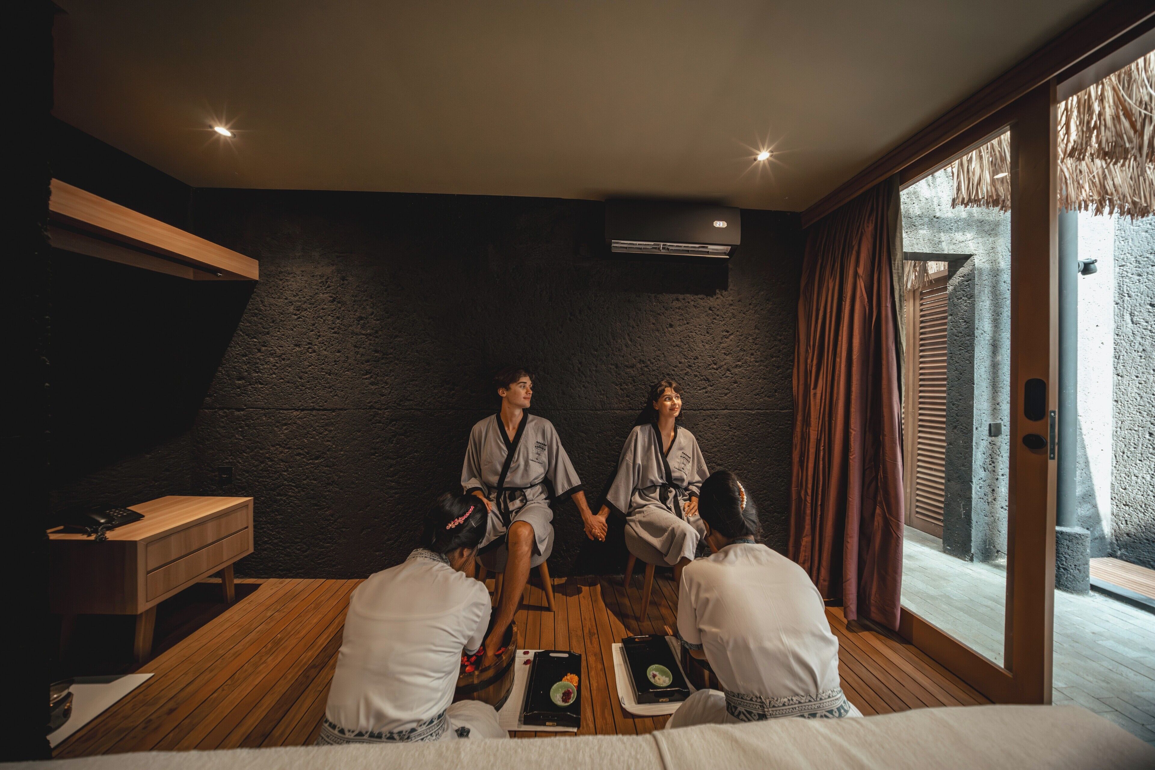 A room with a dark textured wall, a wooden bench, a small wooden table with a tray of food, and a large window with curtains.