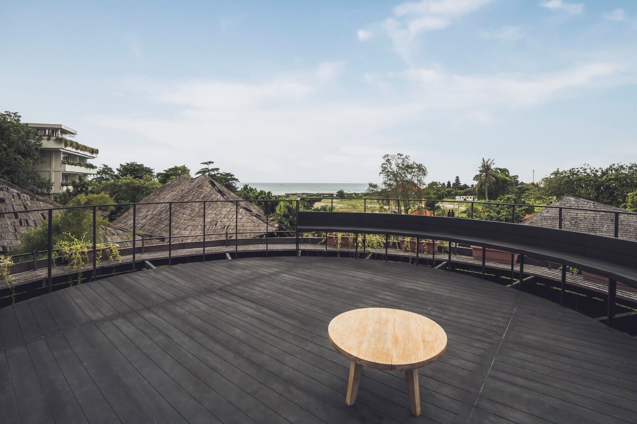 A wooden deck with a round table, surrounded by a black railing, overlooking a beach and greenery.