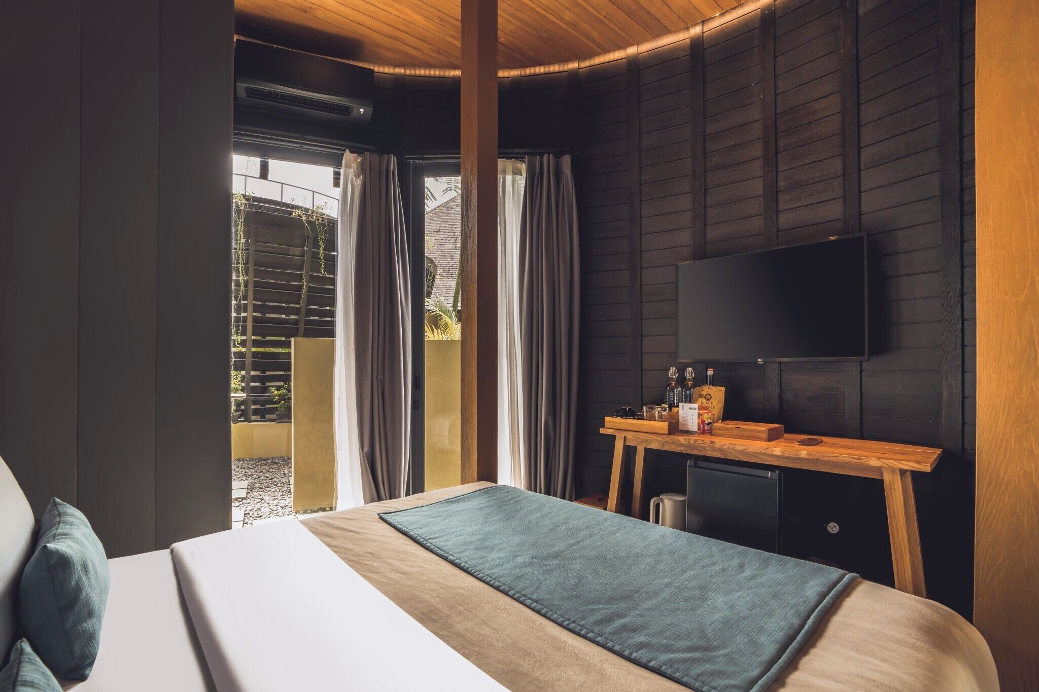 A modern hotel room with a large bed, a wooden desk with a flat-screen TV, and a view of the outdoors through a sliding door.