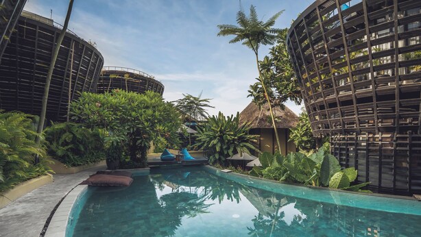 Una piscina con borde de azulejos, rodeada de exuberante vegetación y estructuras arquitectónicas modernas.