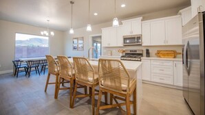 A modern kitchen with white cabinets, a stainless steel refrigerator, a built-in oven, and a dining area with wicker chairs and a table. - Spacious Oasis, Private Pool, Spa & Putting Green (Hurricane)