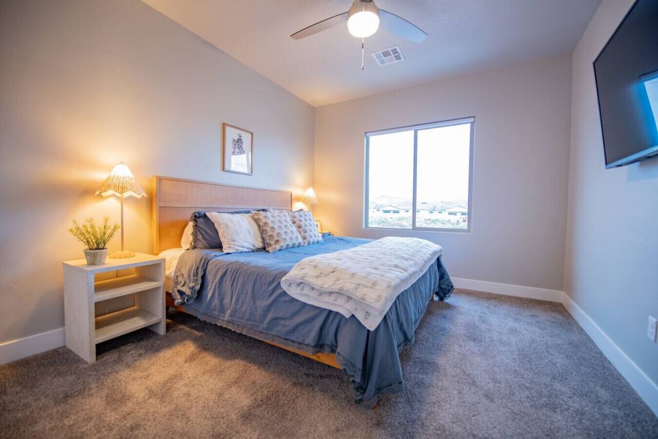 A bedroom with a bed, bedside tables, a ceiling fan, a window, and a TV mounted on the wall.