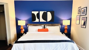 A large bed with a grey headboard, white bedding, and an orange pillow. There are two bedside tables with lamps. The room has a blue accent wall with abstract art and framed pictures. - Mod KING Studio Apartment w/private covered patio (Southfield)