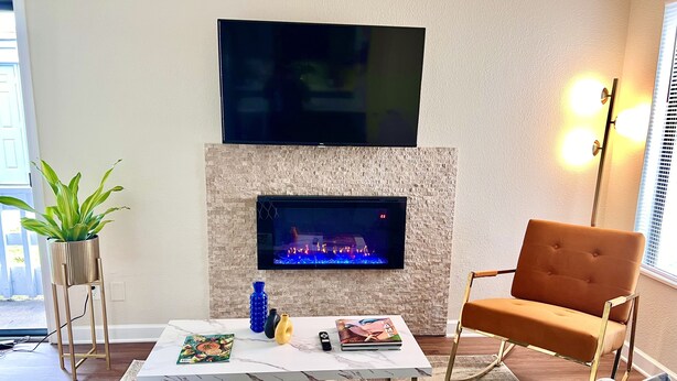 A living room with a flat-screen TV mounted on a textured wall, a fireplace with a TV screen, a coffee table with magazines and a blue vase, a brown armchair, and a potted plant. - Mod KING Studio Apartment w/private covered patio (Southfield)