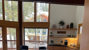 Un amplio comedor con una mesa y bancos de madera, ventanas grandes con vistas a árboles y un edificio, y una zona de cocina con armarios de madera y una encimera.