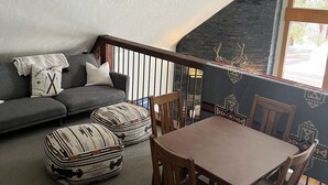 Un cuarto acogedor con un sofá gris, un otomano estampado, una mesa de comedor de madera y sillas. Una escalera conduce a un nivel superior con una pared de ladrillo negro y una ventana.