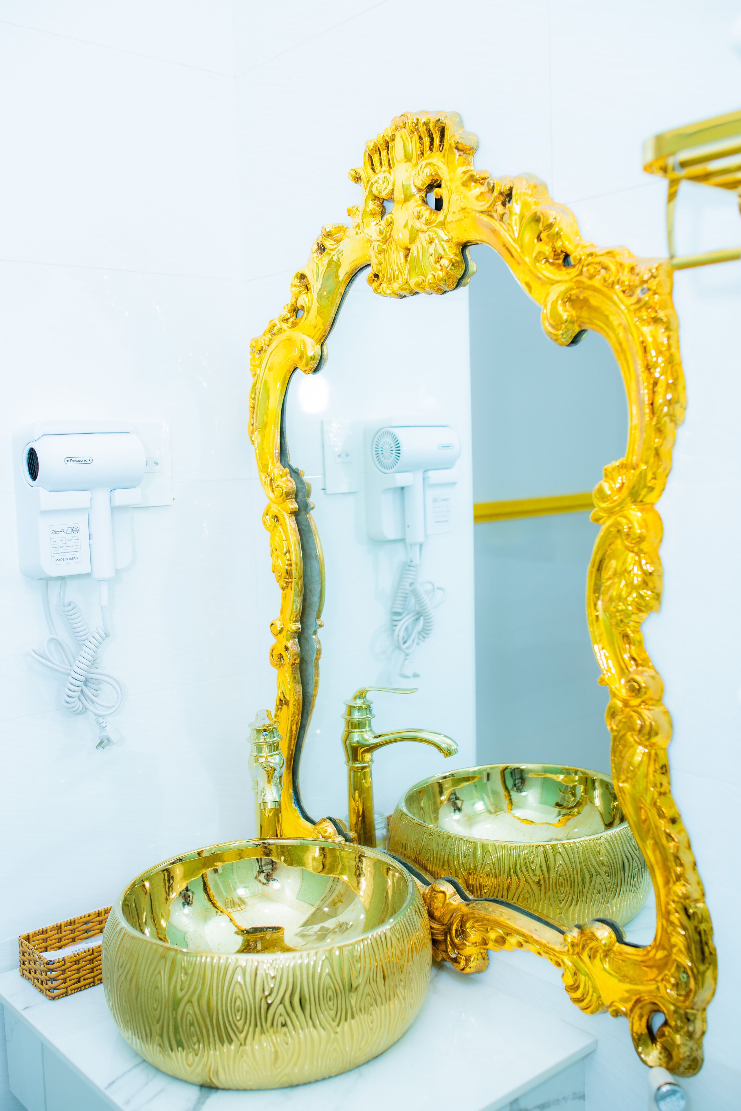 Un baño con un espejo con marco dorado, dos lavamanos con recipientes dorados y una pared blanca con un secador de pelo y dispensador de jabón.