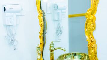 Un baño con un espejo con marco dorado, dos lavamanos con recipientes dorados y una pared blanca con un secador de pelo y dispensador de jabón.