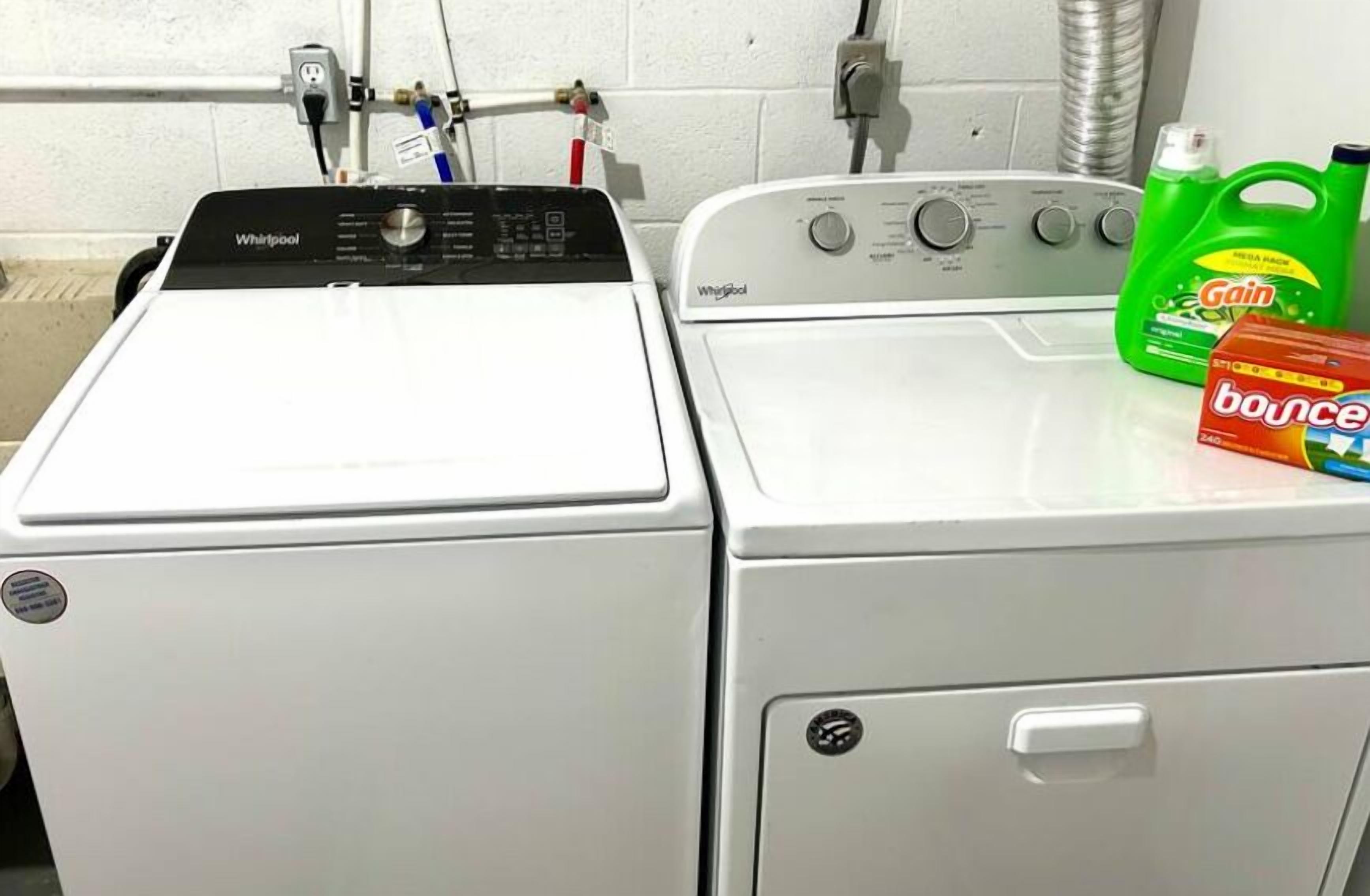 Uma máquina de lavar e secar Whirlpool de abertura frontal, um painel de controle, uma garrafa verde de detergente Gain e uma caixa de amaciante Bounce sobre a secadora.