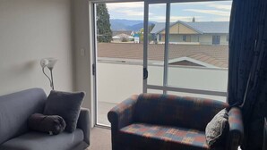Una habitación con un sofá gris, un sillón escocés y un amplio ventanal corredero que da a un balcón con vistas a casas y montañas.