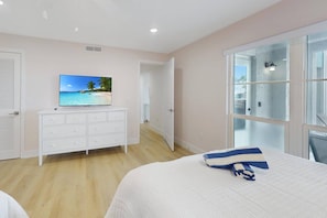 A bedroom with a bed, a dresser, a television displaying a beach scene, and a large window with a view of the outdoors.