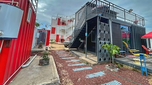 A modern building with a red and white facade, a staircase leading to a balcony, and a small garden area with plants and seating.